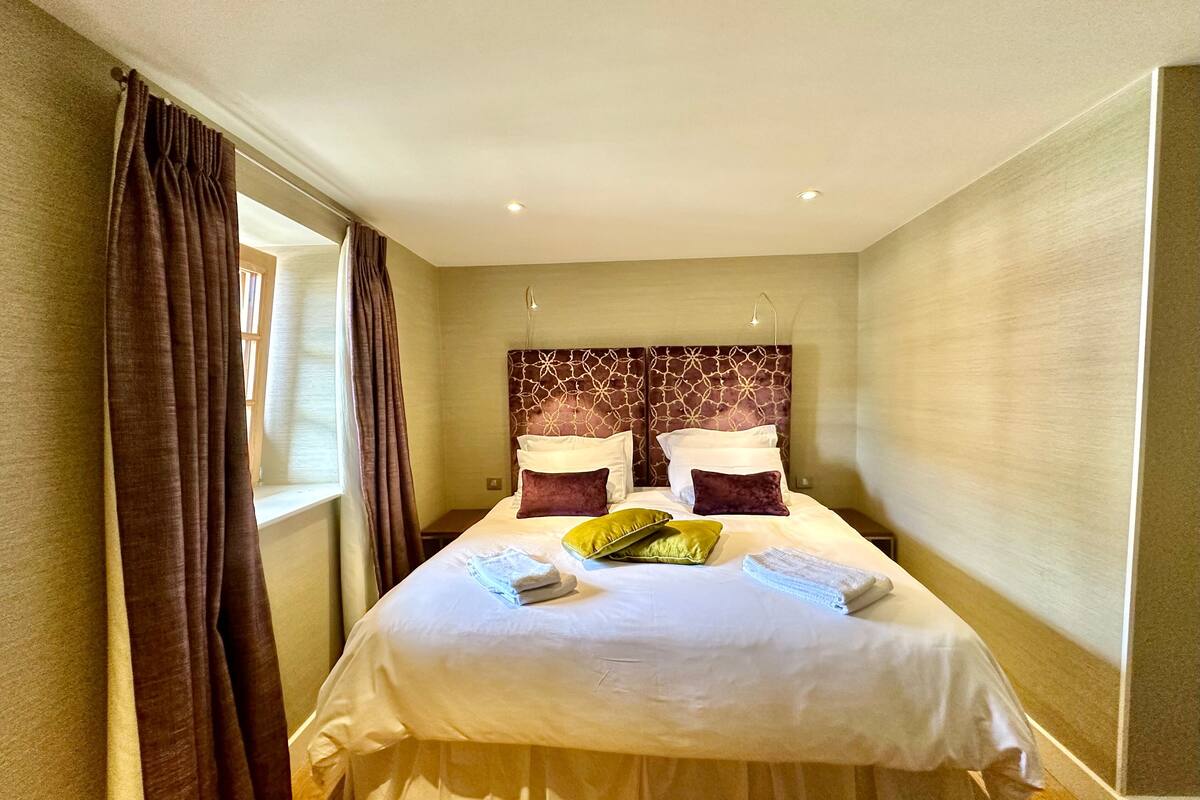 A neatly made bed with white linens and multiple pillows, a window with curtains, and a headboard with a geometric pattern.