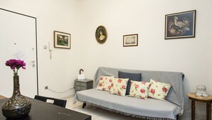A small, tidy room with a sofa, a table with a vase of flowers, and a few framed pictures on the wall.