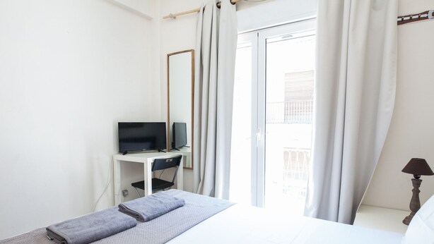 A hotel room with a bed, a desk with a TV, a window with curtains, and a lamp.