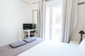 A hotel room with a bed, a desk with a TV, a window with curtains, and a lamp.