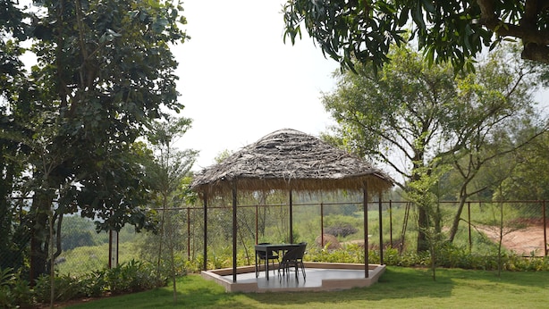 A thatched-roof gazebo with a table and chairs in a grassy area surrounded by trees.