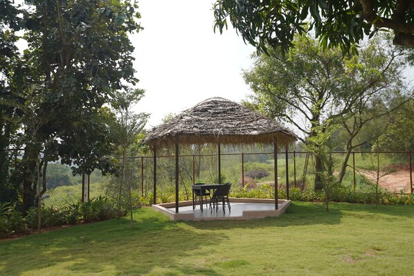 A thatched-roof gazebo with a table and chairs in a grassy area surrounded by trees.