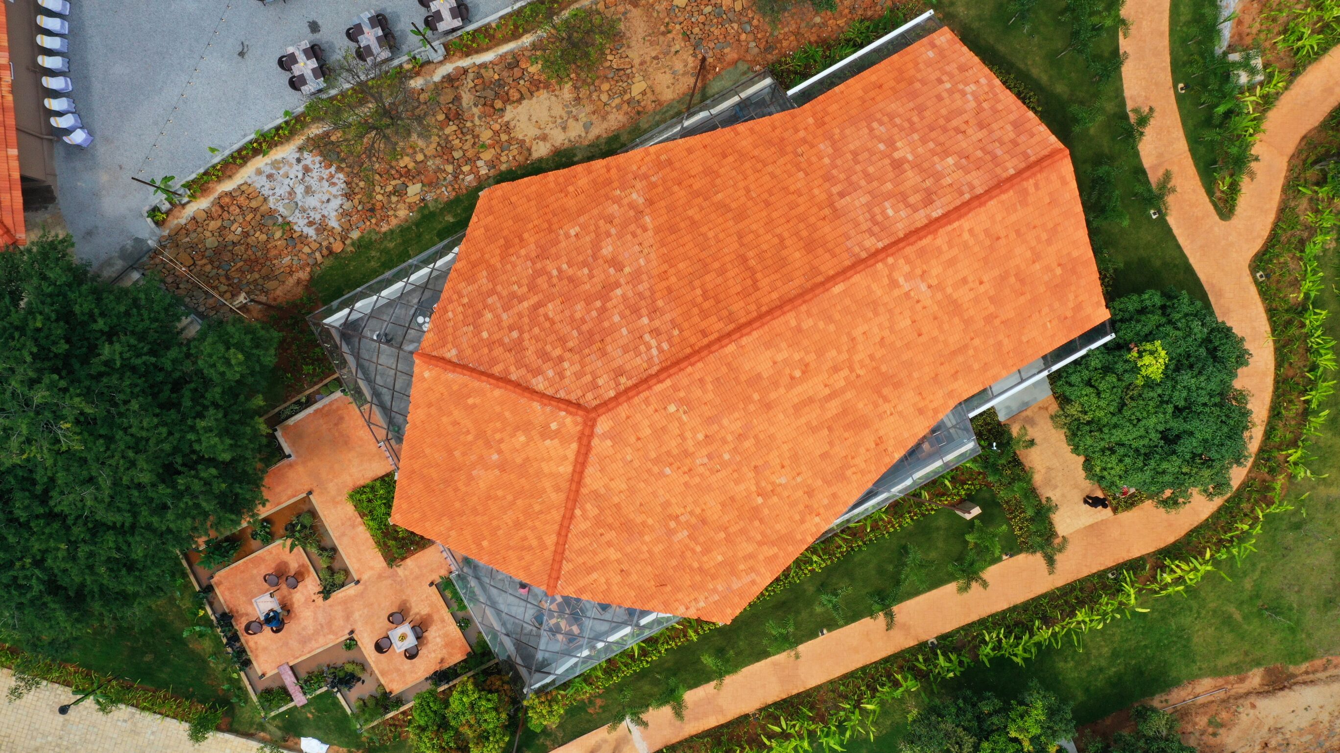 An aerial view of a building with a large orange roof, surrounded by greenery and a paved area with seating.