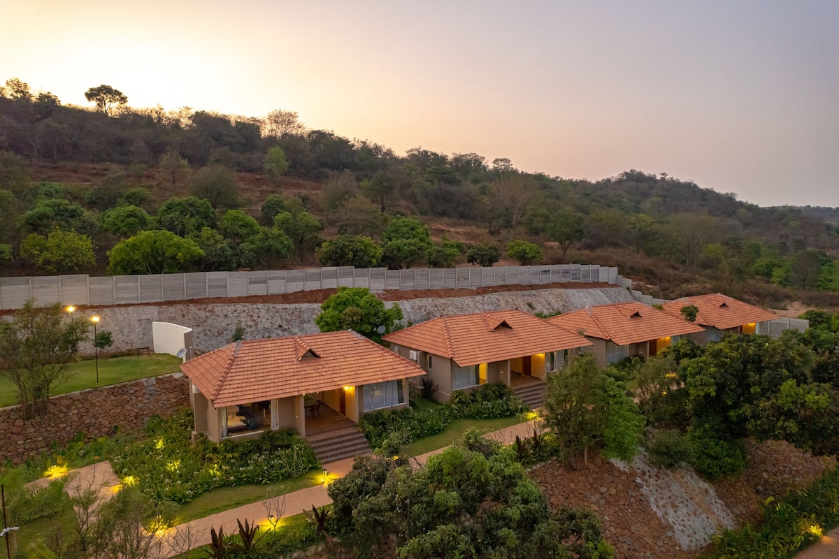 A resort with multiple bungalow-style buildings, each with a tiled roof and a small porch. The resort is surrounded by lush greenery and hills in the background.