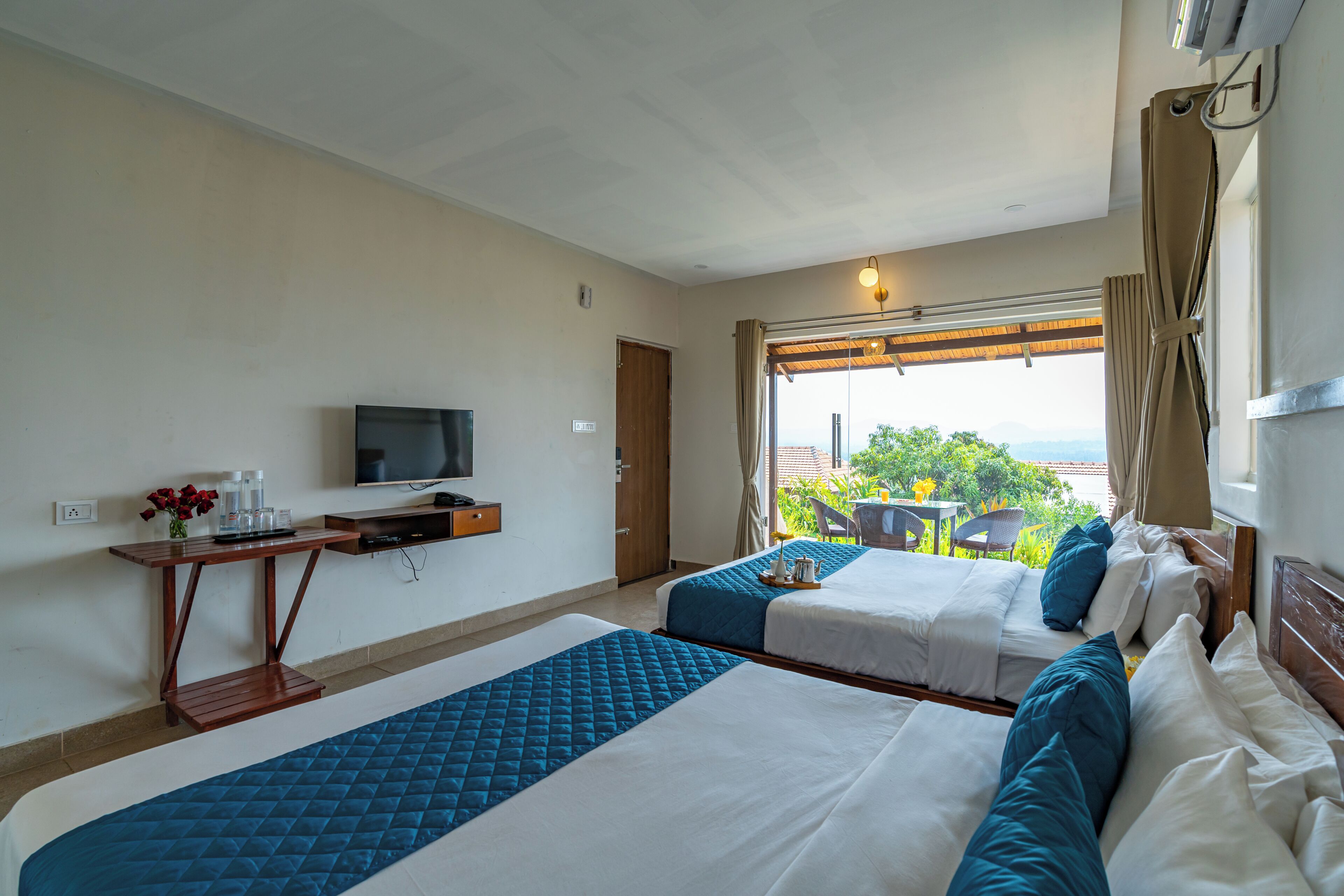 Habitación de hotel con dos camas, un televisor, una mesita con un jarrón de flores y vistas al exterior.