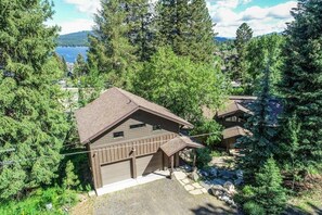A cabin with a sloped roof, a garage, and a covered porch, surrounded by dense forest and a lake view.