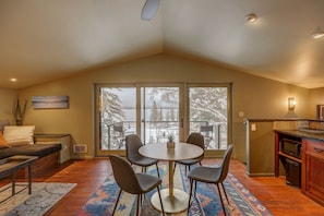 A cozy living room with a sofa, a coffee table, and chairs. There is a large window with a view of trees and a snowy landscape. - Mackinaw Minnow - Payette Lake Views - Downtown McCall Apt- Walk to Lake, Restaurants, Bars, Marina (McCall)