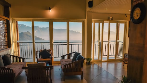 A room with a large sliding glass door leading to a balcony with a view of mountains. There are wooden chairs and a small table in the room. - Chill Berg Resort - Bodimettu (Bodinayakanur)
