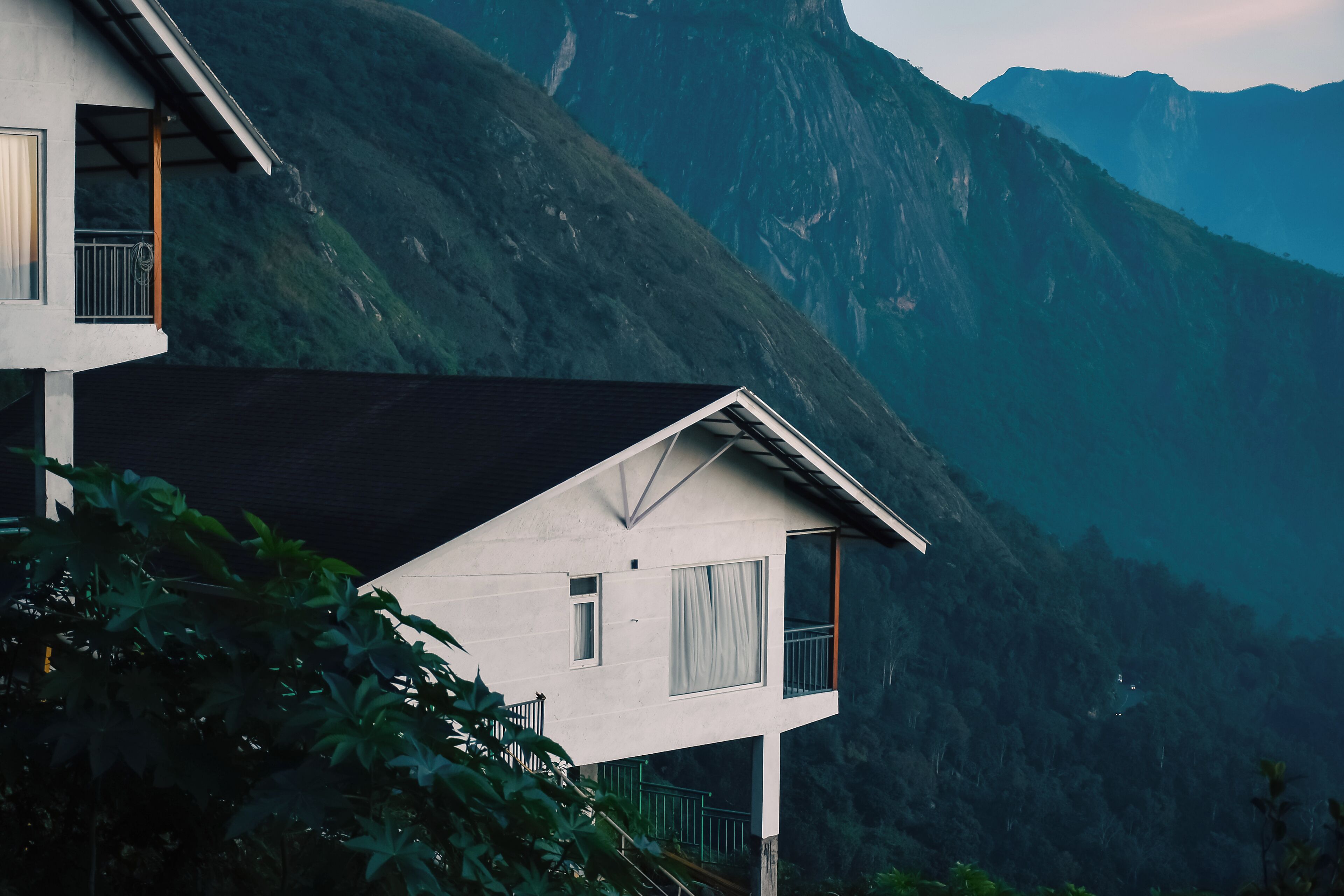 A modern two-story house with a balcony, surrounded by lush greenery and mountains.