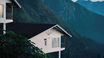 A modern two-story house with a balcony, surrounded by lush greenery and mountains.