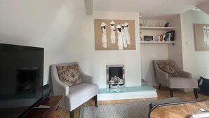 A modern living room with a flat-screen TV, two armchairs, a glass coffee table, a fireplace, and a bookshelf. - Modern Top Floor 2BD Flat - Kensington (London)