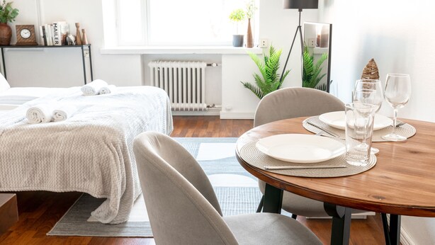 A hotel room with a neatly made bed, a dining table set with plates and glasses, a radiator, a potted plant, and a floor lamp. - 2ndhomes Cozy Apartment with Park View (Helsinki)