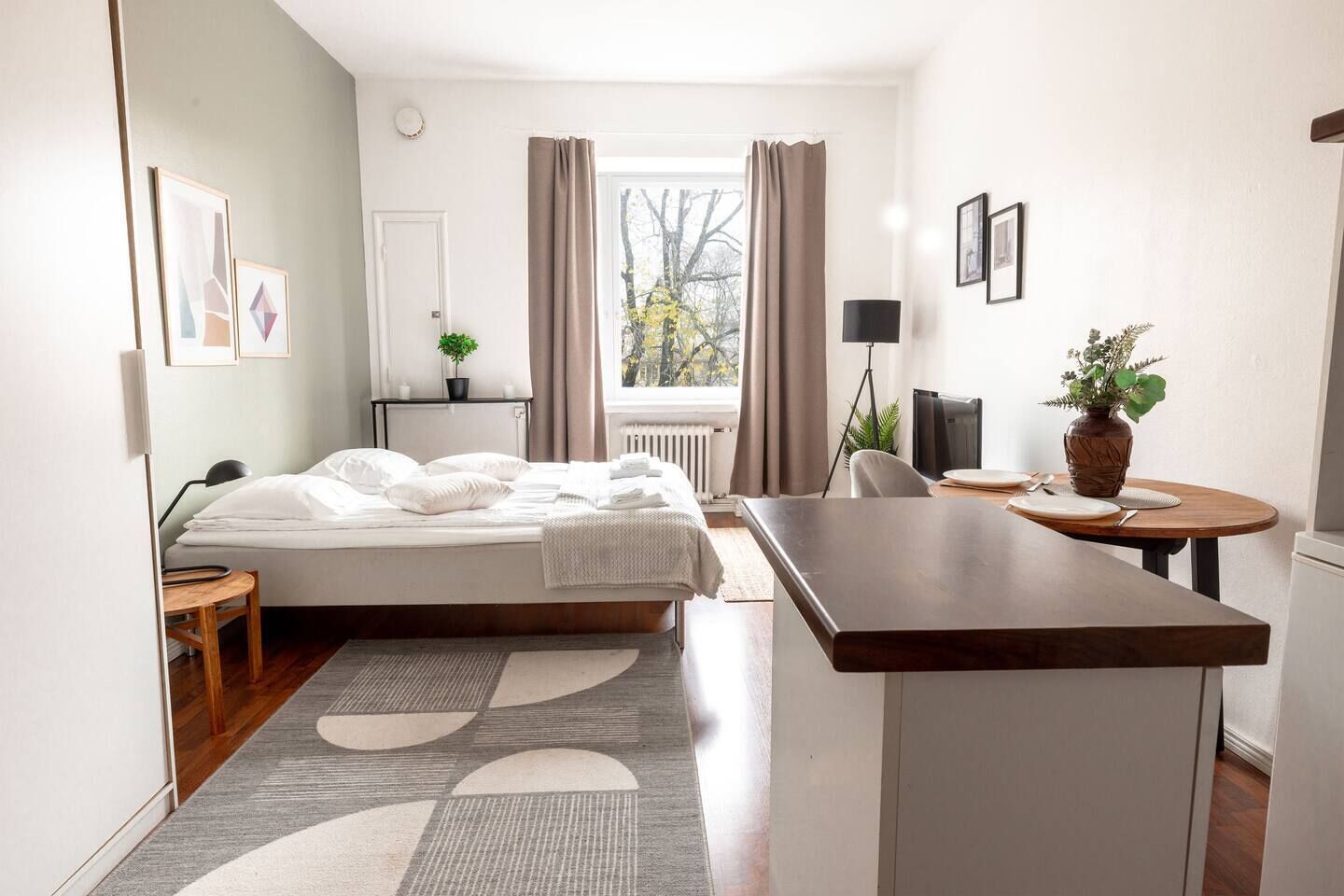 A modern hotel room with a bed, a desk, a chair, a small table, a potted plant, and a window with curtains.