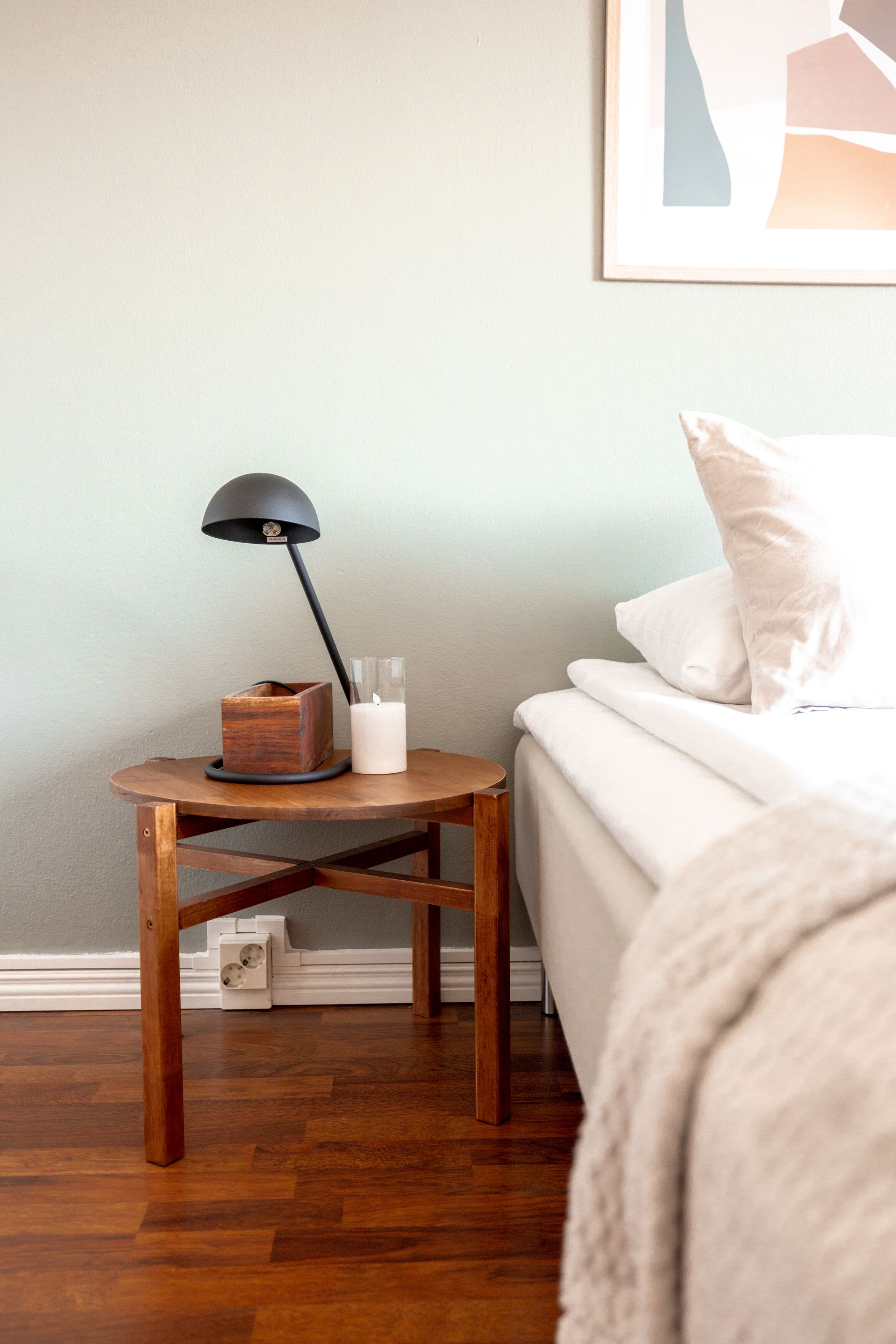 A neatly made bed with white bedding, a small wooden nightstand with a black lamp, and a framed picture on the wall.