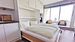 Una habitación de hotel moderna con una cama de madera, armarios blancos, una kitchenette pequeña y un balcón con vistas.