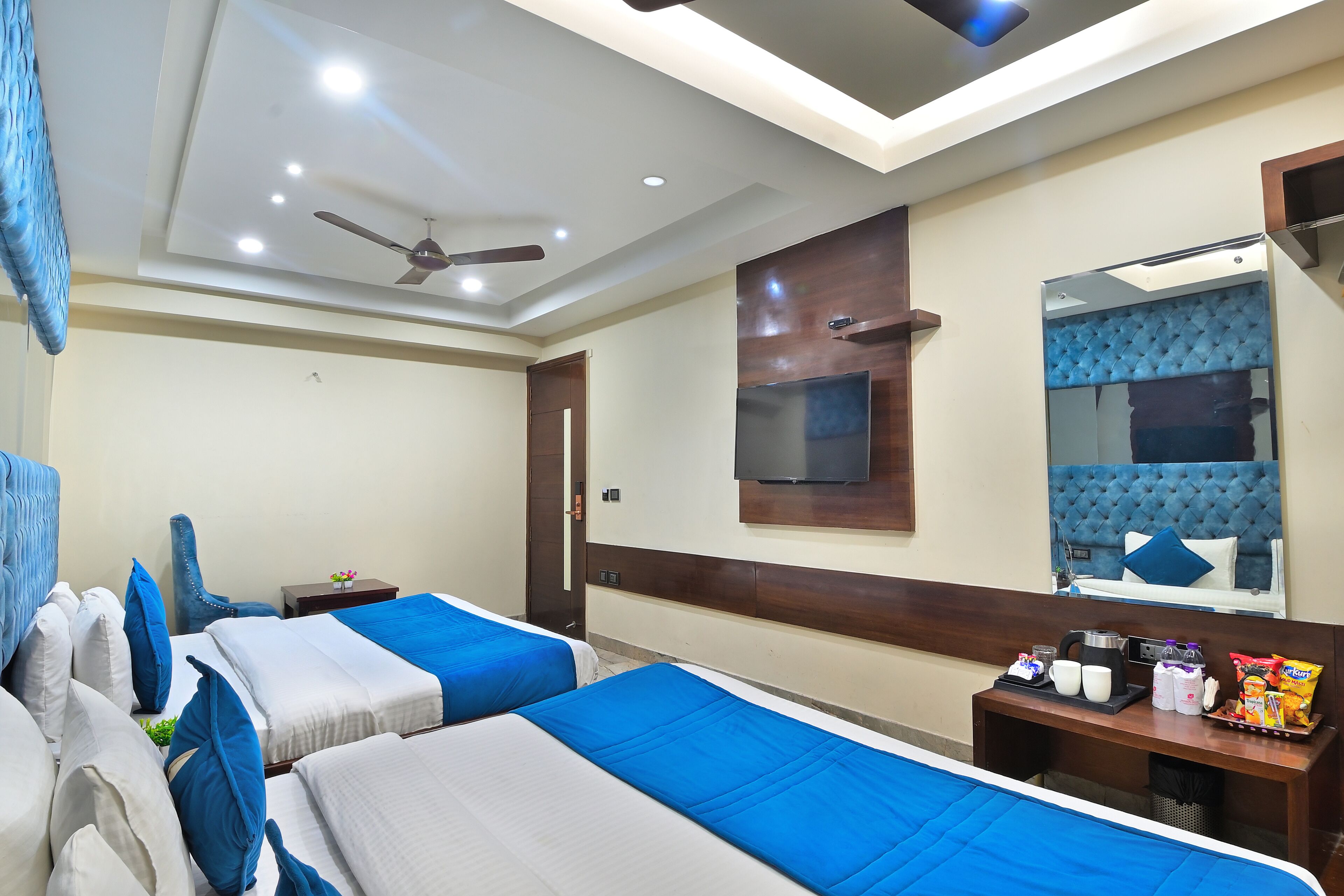 A hotel room with two beds, a TV, a mirror, a ceiling fan, and a small table with snacks and drinks.