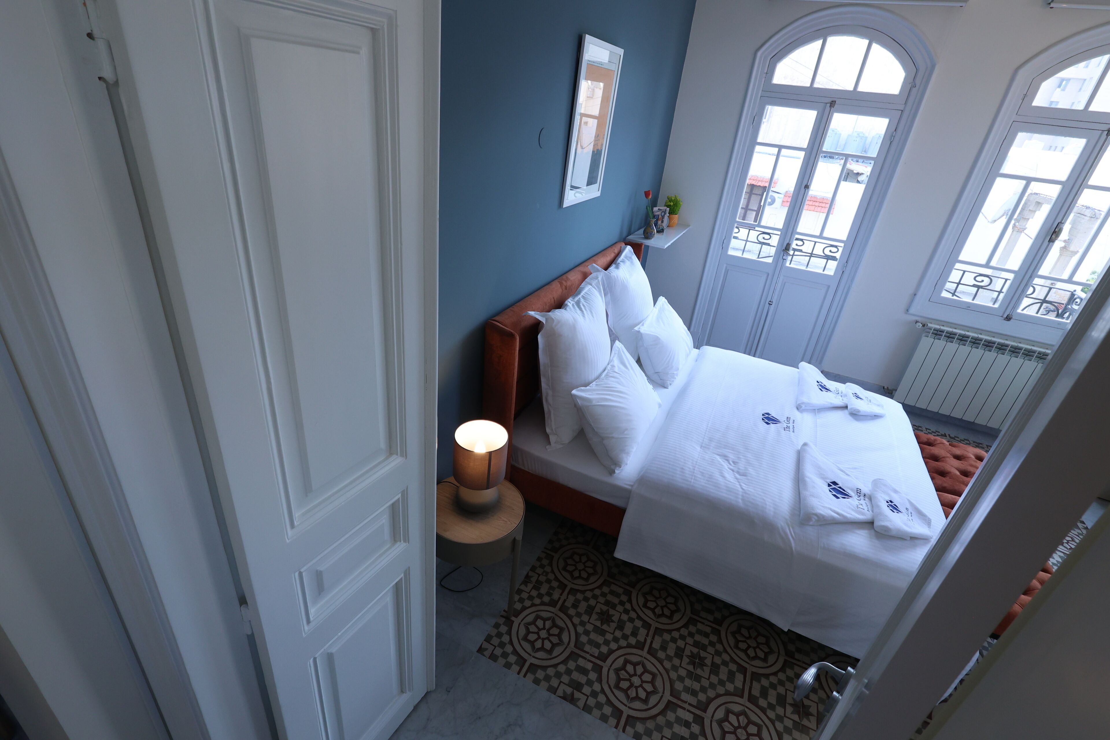 Habitación de hotel con cama, mesita de noche con lámpara, alfombra estampada y vistas al exterior a través de amplias ventanas.
