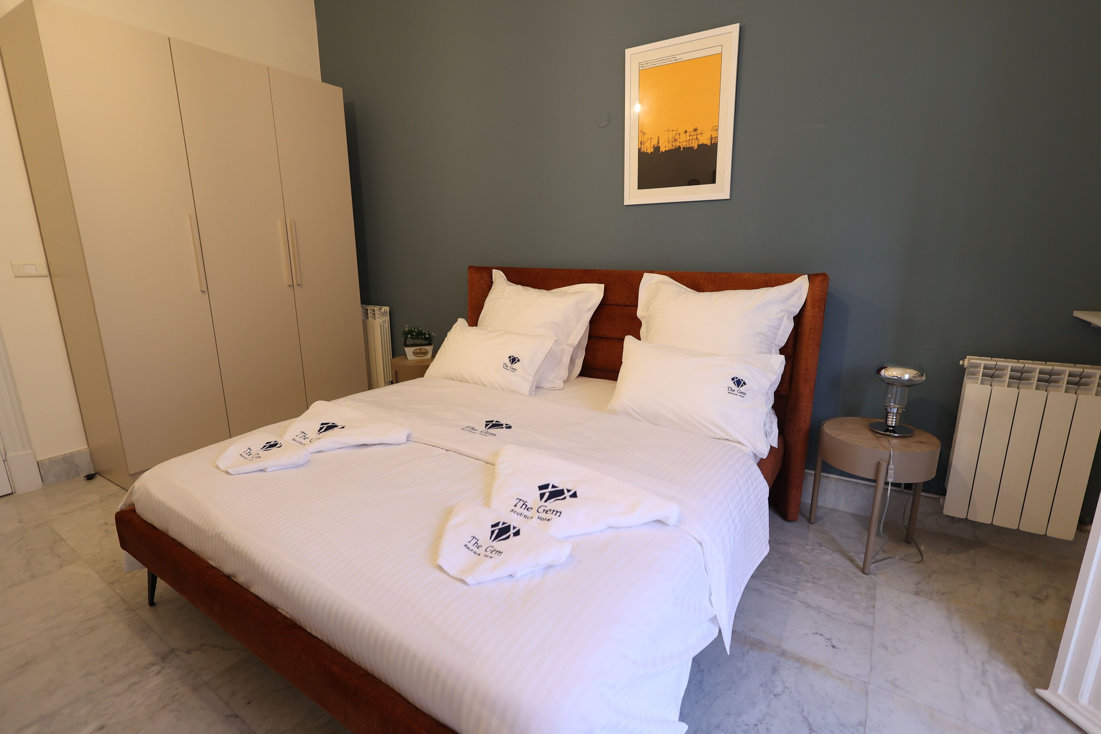 A neatly made bed with white linens and towels, a wooden headboard, a small bedside table with a lamp, and a radiator on the wall.