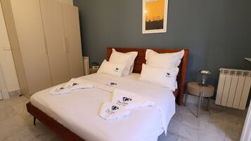 A neatly made bed with white linens and towels, a wooden headboard, a small bedside table with a lamp, and a radiator on the wall.
