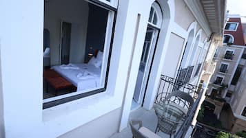Un balcón con una silla de mimbre, una cama con ropa blanca y vista a la calle.