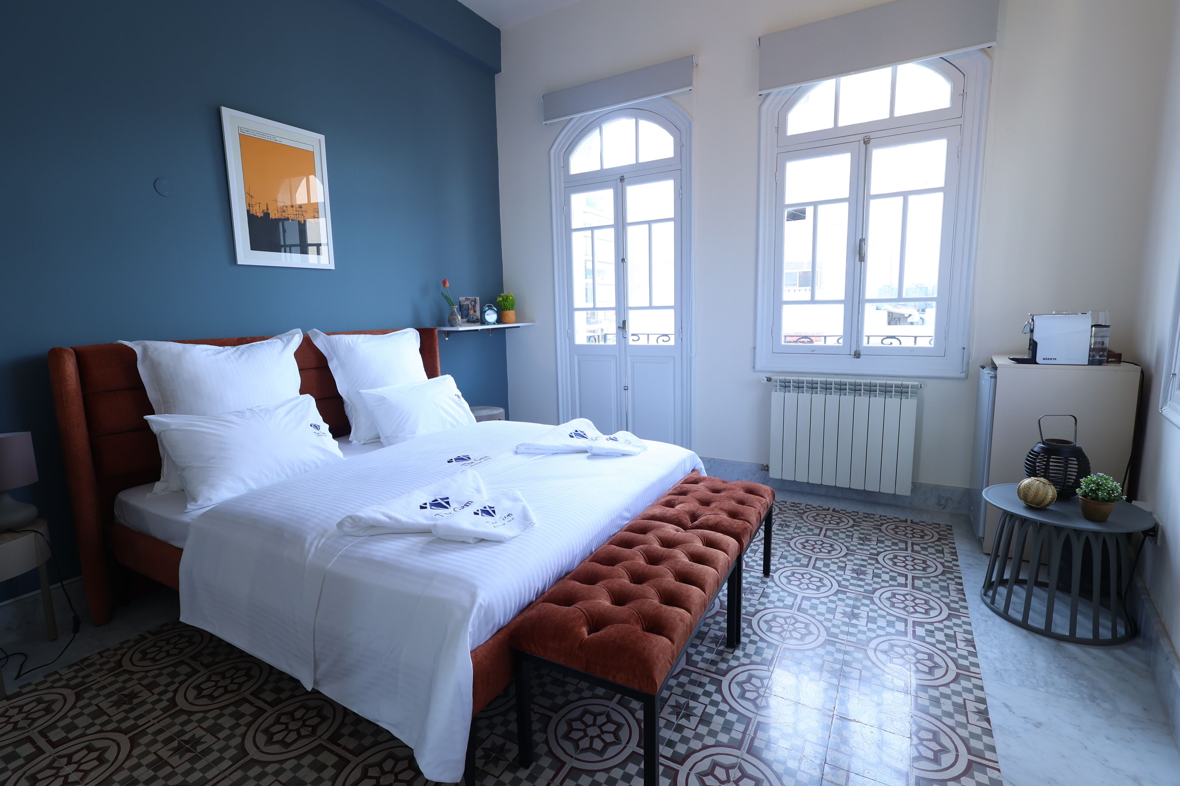 A neatly made bed with white linens, a brown bench, and a patterned floor.