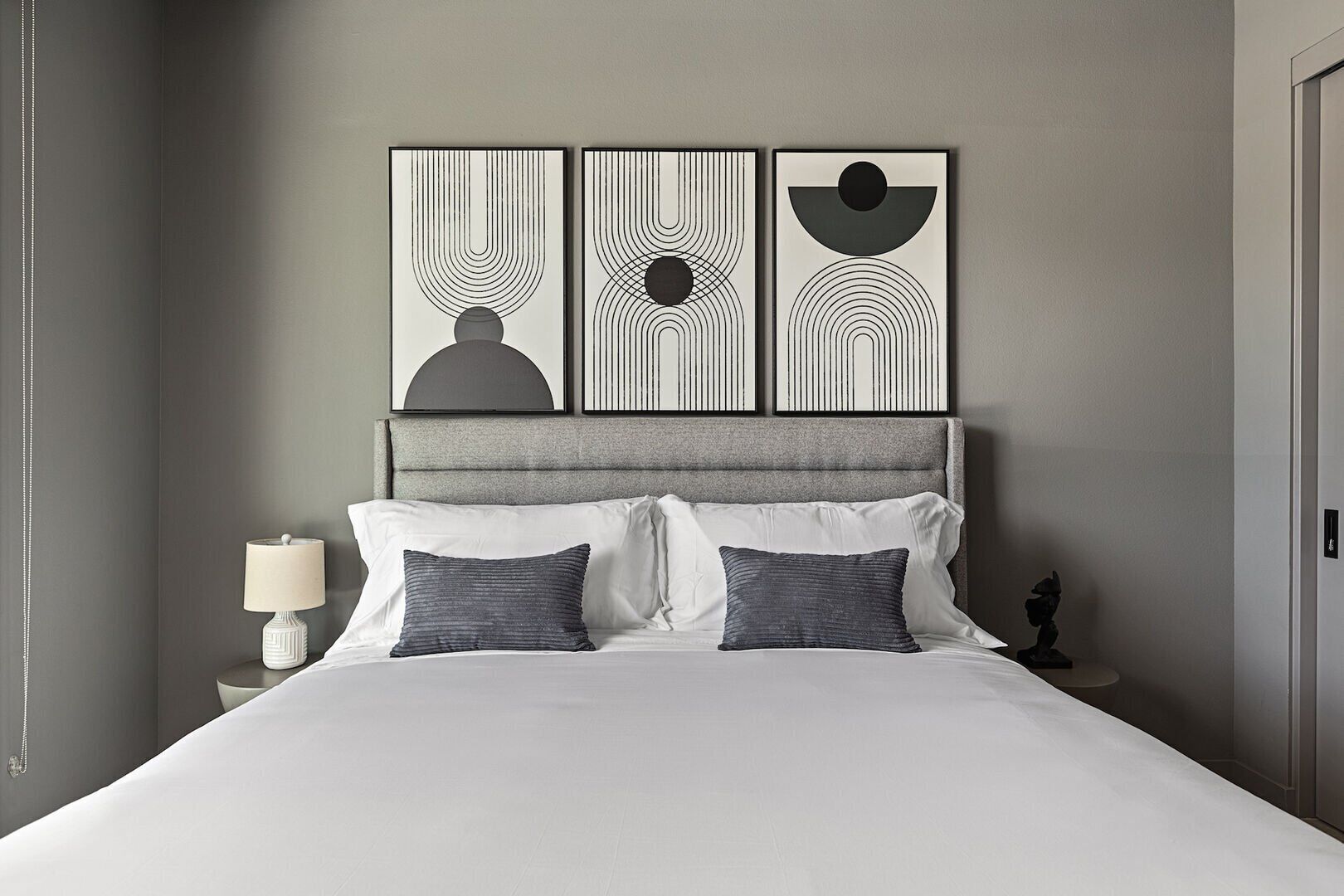 A neatly made bed with white linens and two gray pillows, a gray upholstered headboard, and a nightstand with a white lamp.