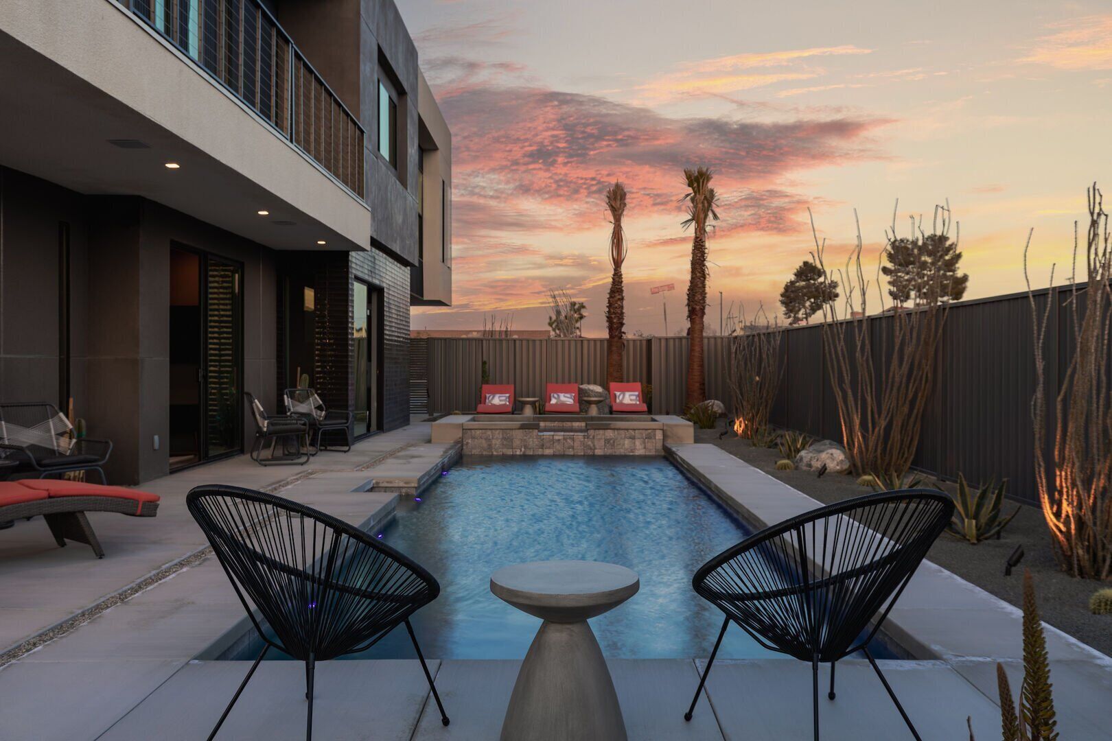A modern outdoor pool area with two lounge chairs, a fire pit, and a patio with a view of the sunset.