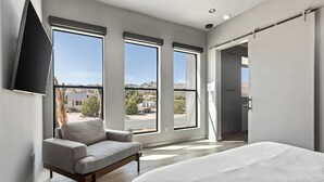 Un dormitorio moderno con una cama grande, un sillón gris y tres ventanas amplias que ofrecen vistas al exterior.