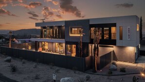 Una casa moderna de dos pisos con un balcón, ventanas amplias y un paisaje desértico.
