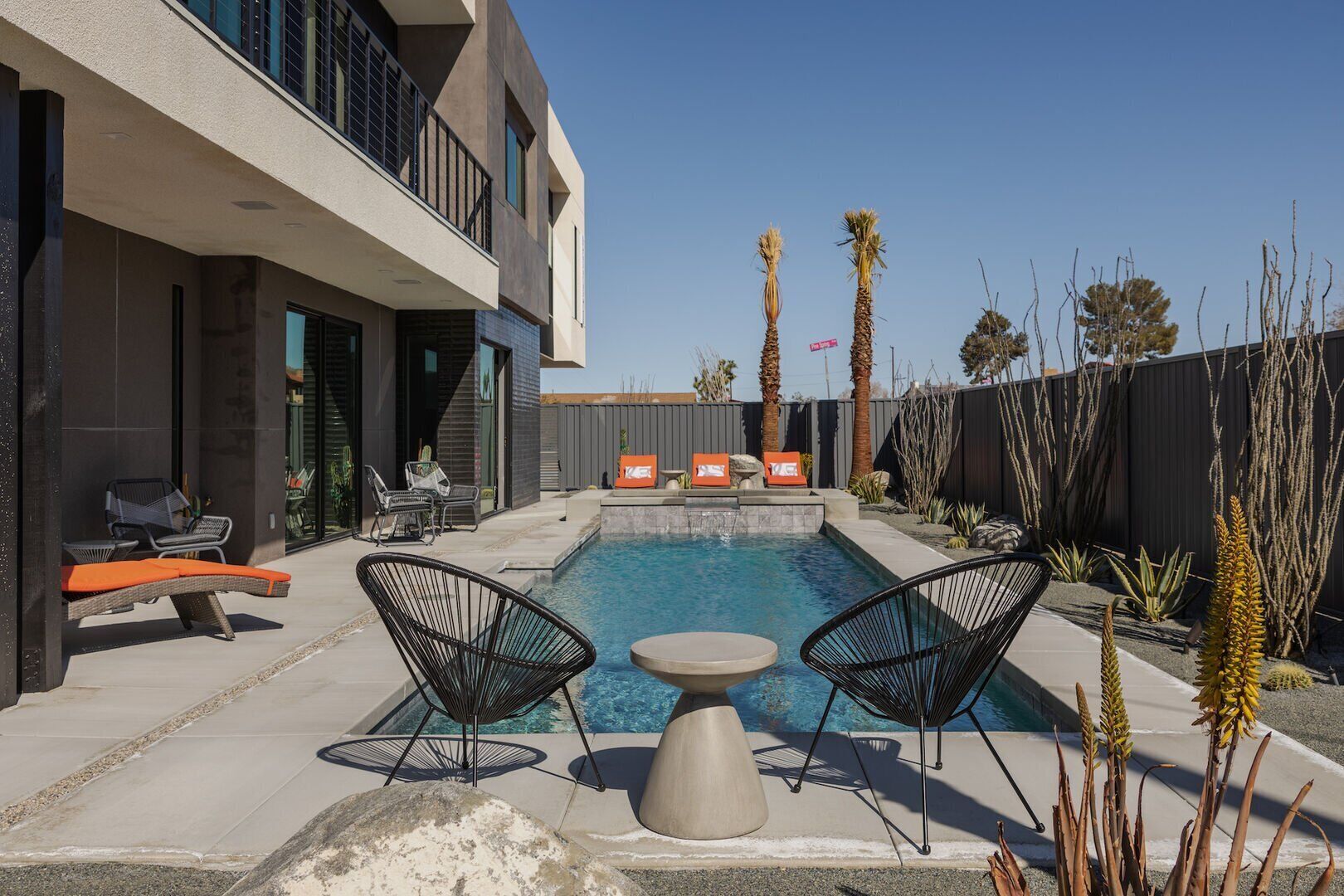 Un espace piscine extérieur moderne, comprenant deux chaises longues, une petite table et un coin salon avec des coussins orange.