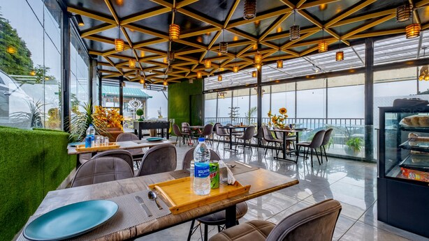 A modern restaurant with a high ceiling, geometric design, and ample natural light. Tables are set with plates, cutlery, and a bottle of water. - Le Masrana Inn (Dehradun)