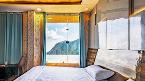 A hotel room with a large bed, a desk, and a view of mountains through a window. - Le Masrana Inn (Dehradun)