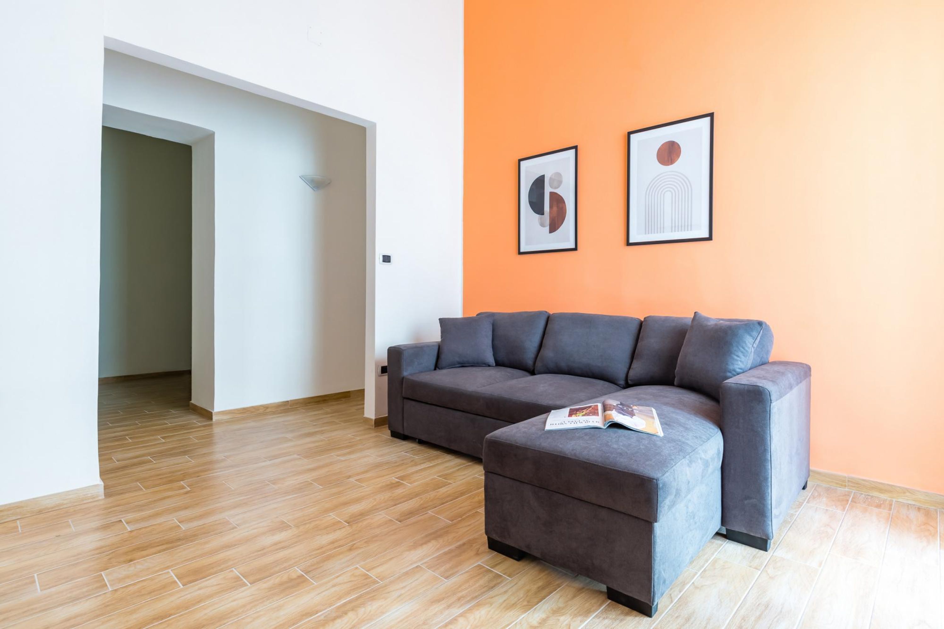 A modern living room with a grey sofa and ottoman, a wooden floor, and two framed artworks on an orange wall.