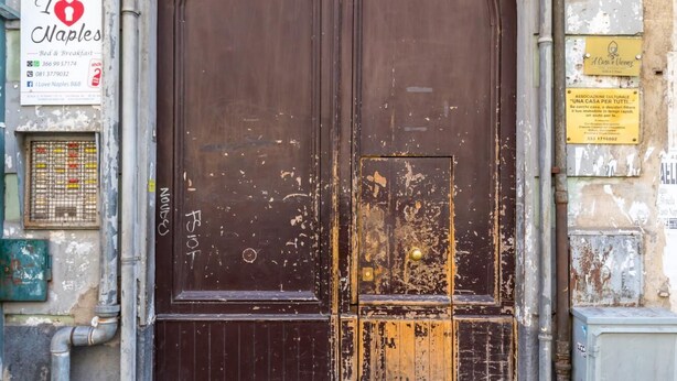A large, arched wooden door with visible wear and tear, set in a weathered building facade with various signs and utility boxes. - Central Station Family Apt by Wonderful Italy (Naples)