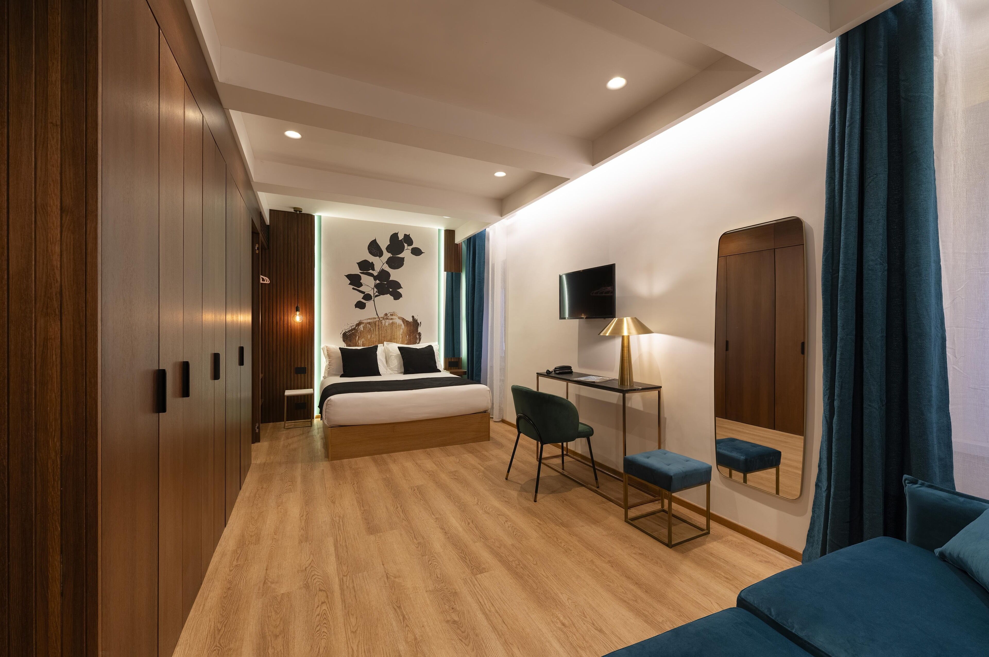 A modern hotel room with a wooden floor, a bed with a headboard, a desk with a chair, a TV, and a mirror.