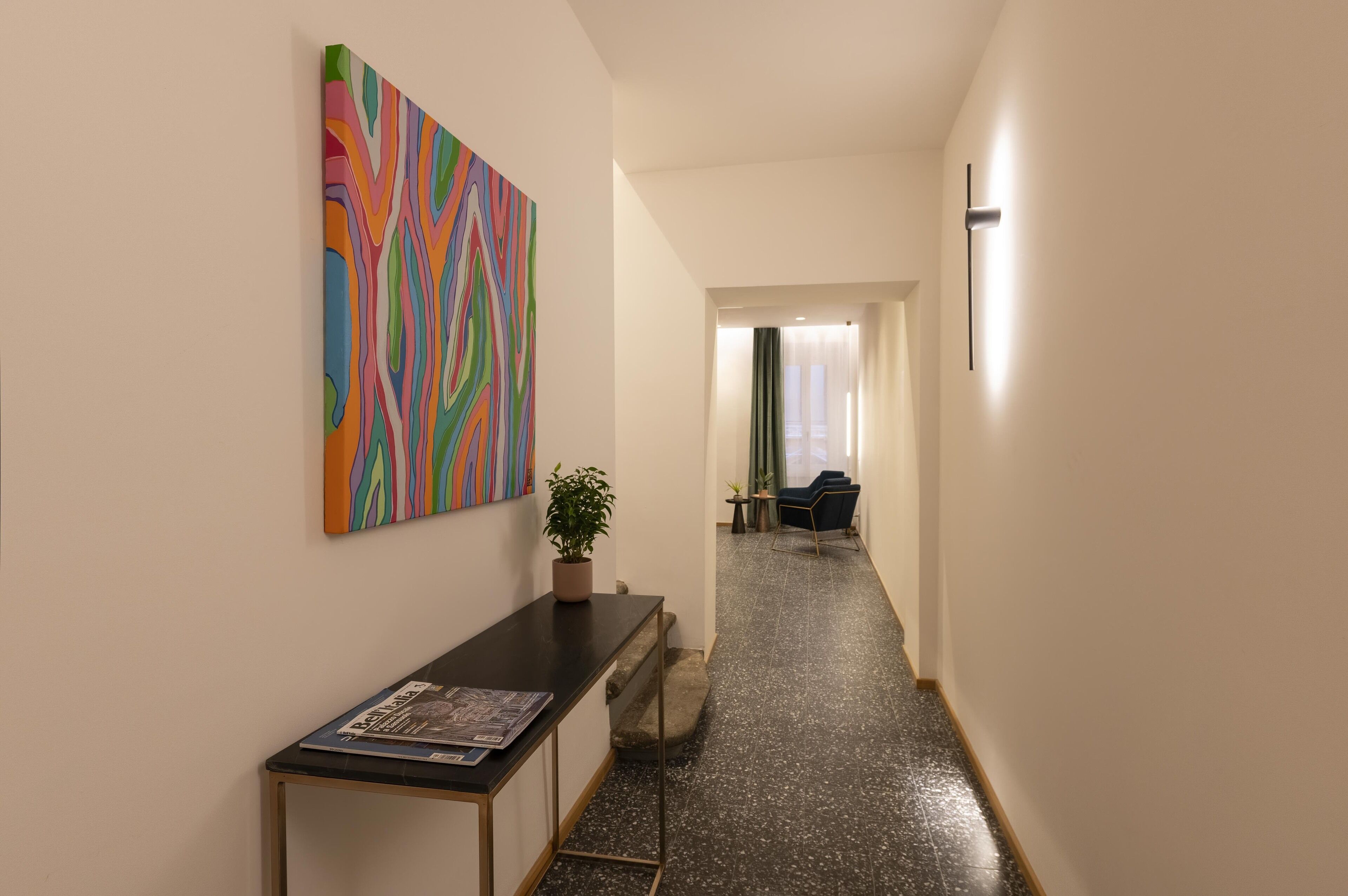 A hallway with a reception desk, a colorful abstract painting, a potted plant, and a seating area with a chair and table.
