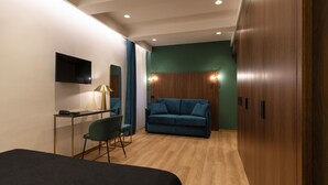 A modern hotel room with a wooden floor, a blue sofa, a desk with a lamp, a TV mounted on the wall, and a wardrobe. - Palazzetto Symphony (Rome)