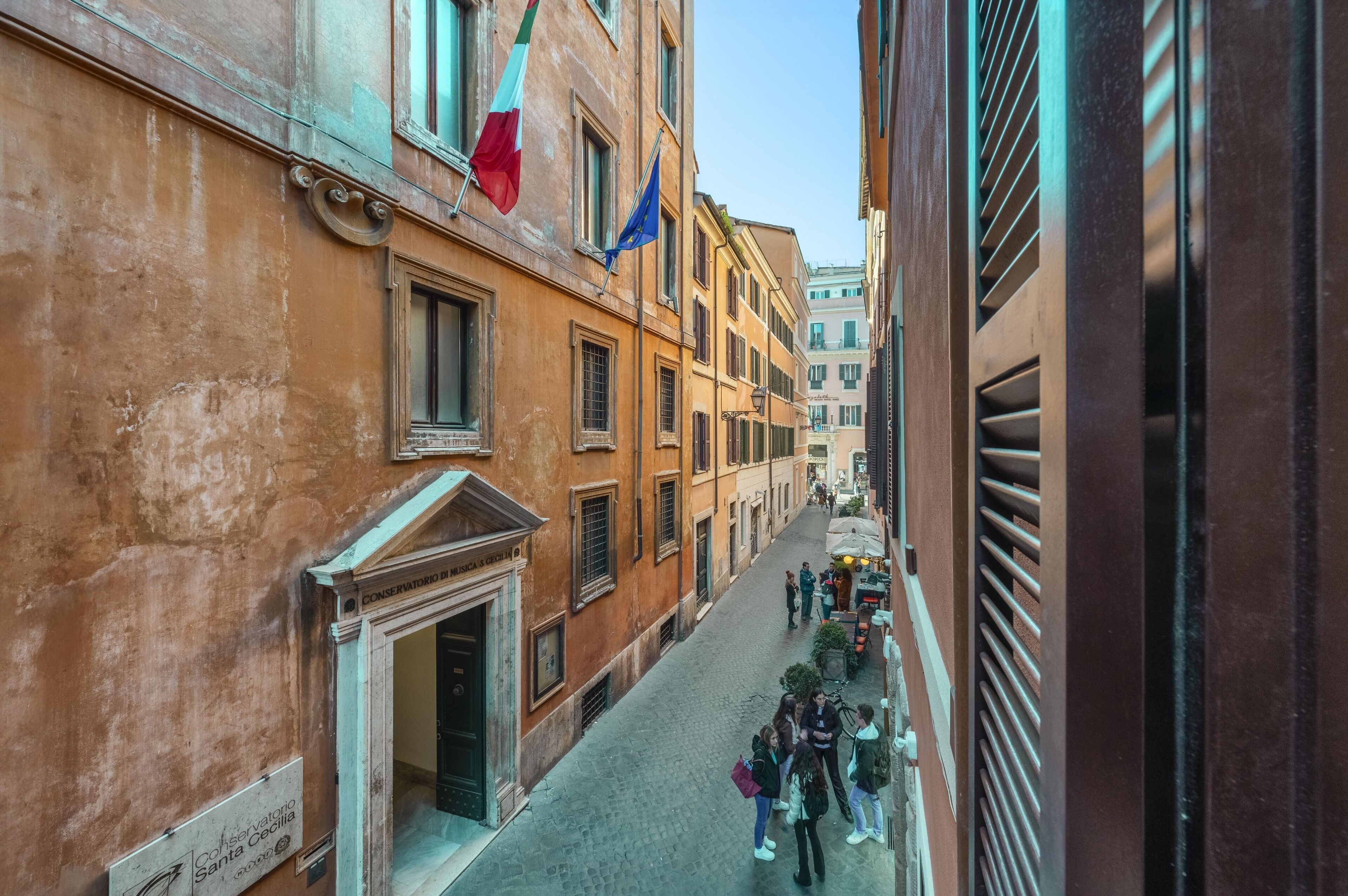 A narrow cobblestone street lined with historic buildings, a flag, and a sign for the Conservatorio di Santa Cecilia.