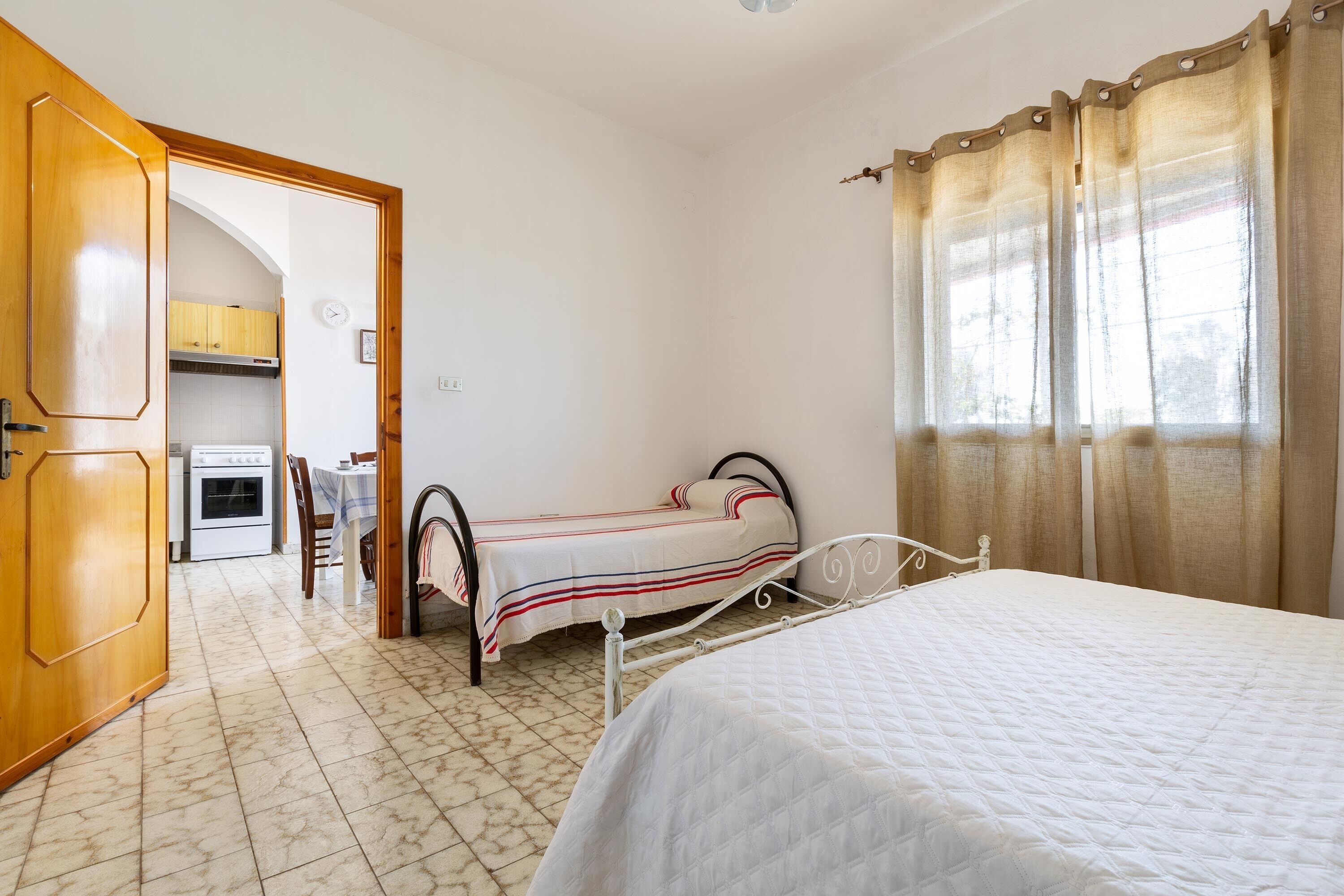 Un dormitorio con una cama, una mesita y una silla. Se ve un área de cocina a través de una puerta abierta.