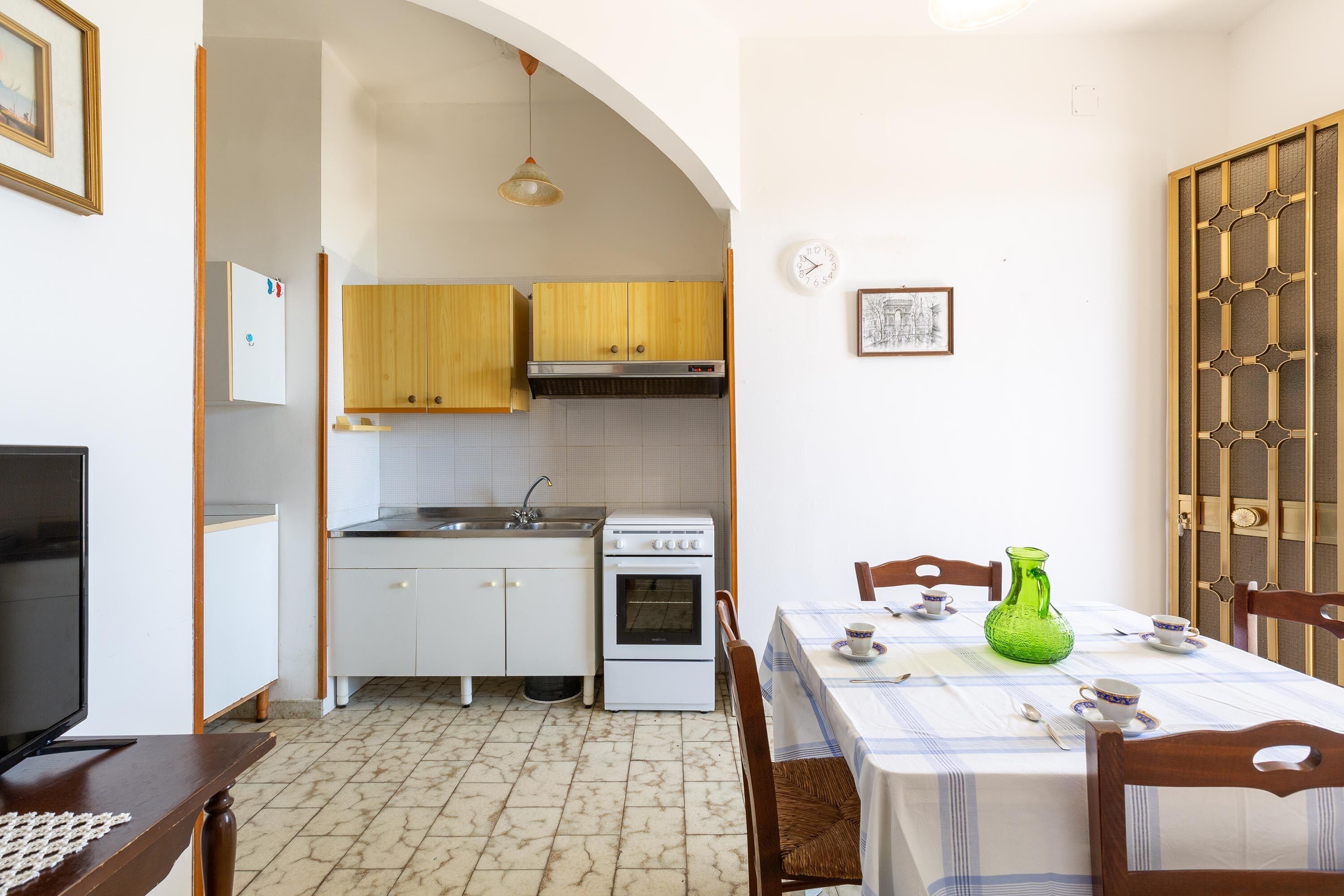 A compact kitchen with white cabinets, a stove, and a sink. There is a dining table with chairs and a green vase.