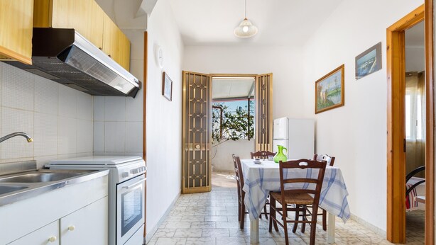 A kitchen with a stove, sink, and cabinets, a dining table with chairs, and a view through a door to an outdoor area. - 3309 Villetta Mare Fuori - Appartamento Piccolo by Barbarhouse (Porto Cesareo)