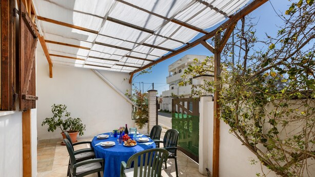 A covered patio with a table set for two, surrounded by chairs, a potted plant, and a view of buildings in the background. - 3310 Villetta Mare Fuori - Appartamento Grande by Barbarhouse (Porto Cesareo)