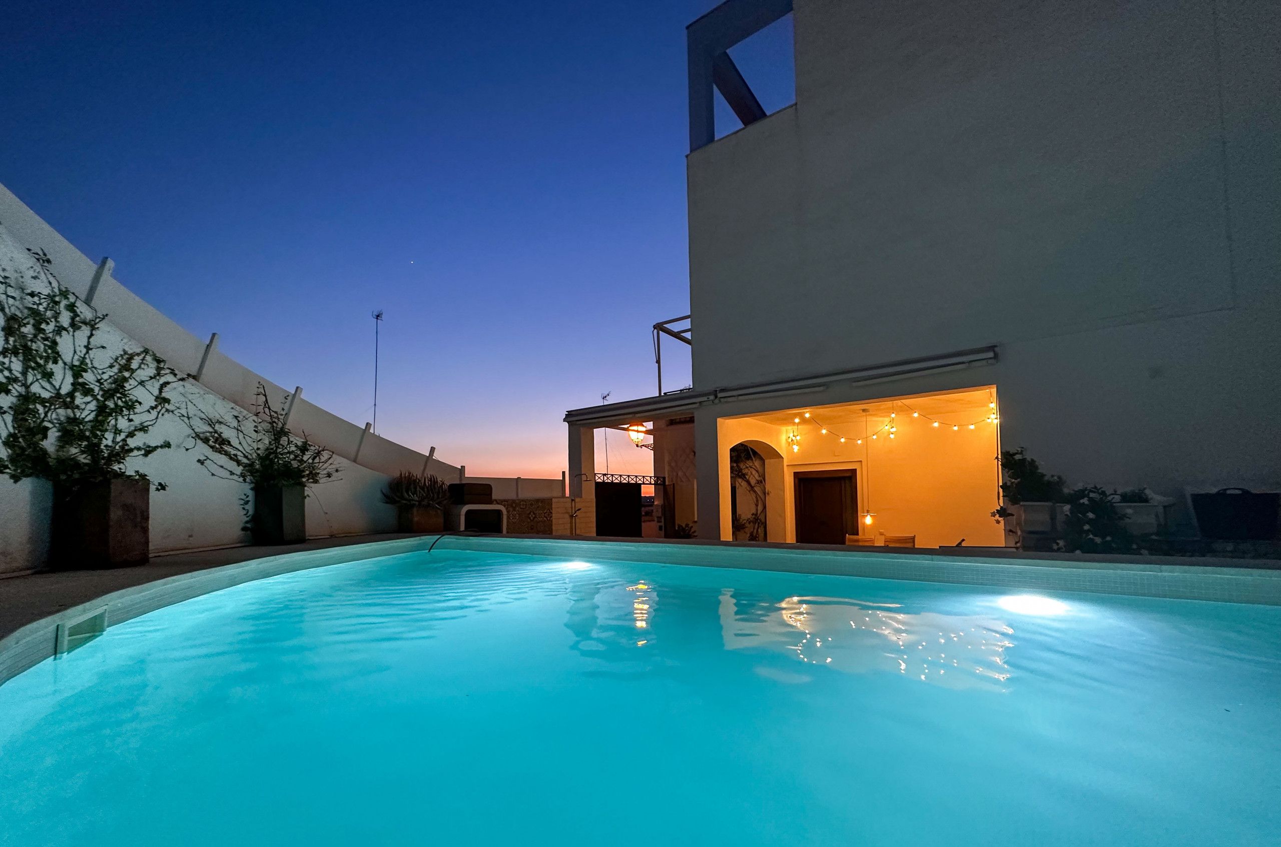 A swimming pool with a covered area and string lights, surrounded by a white building and potted plants.