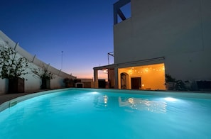 Piscina con zona techada y luces colgantes, rodeada de un edificio blanco y plantas en macetas.