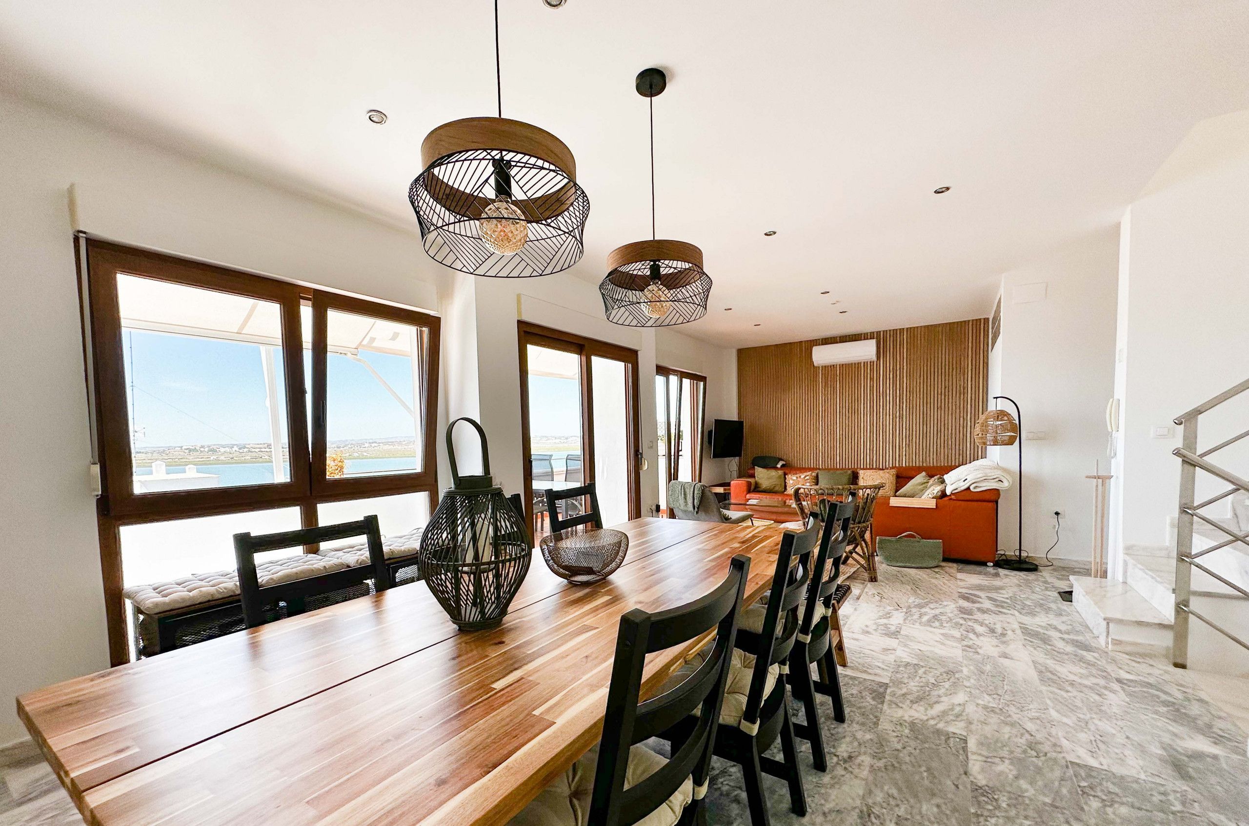 A modern dining area with a wooden table and chairs, large windows, and hanging pendant lights.