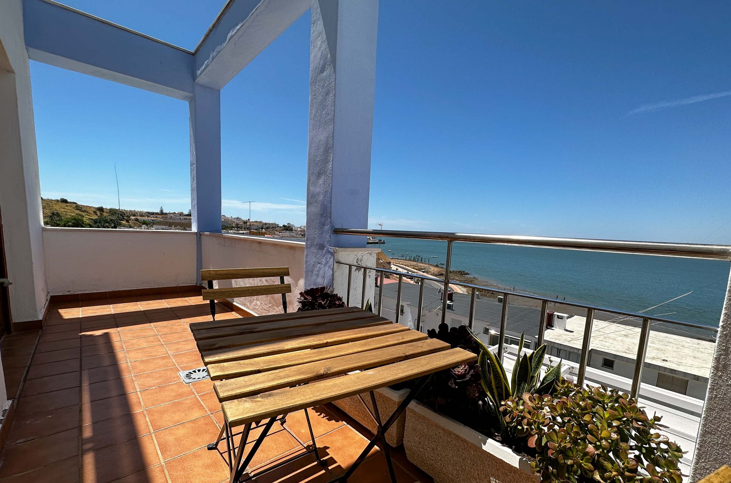 A balcony with a wooden table and chairs, a potted plant, and a view of the sea.