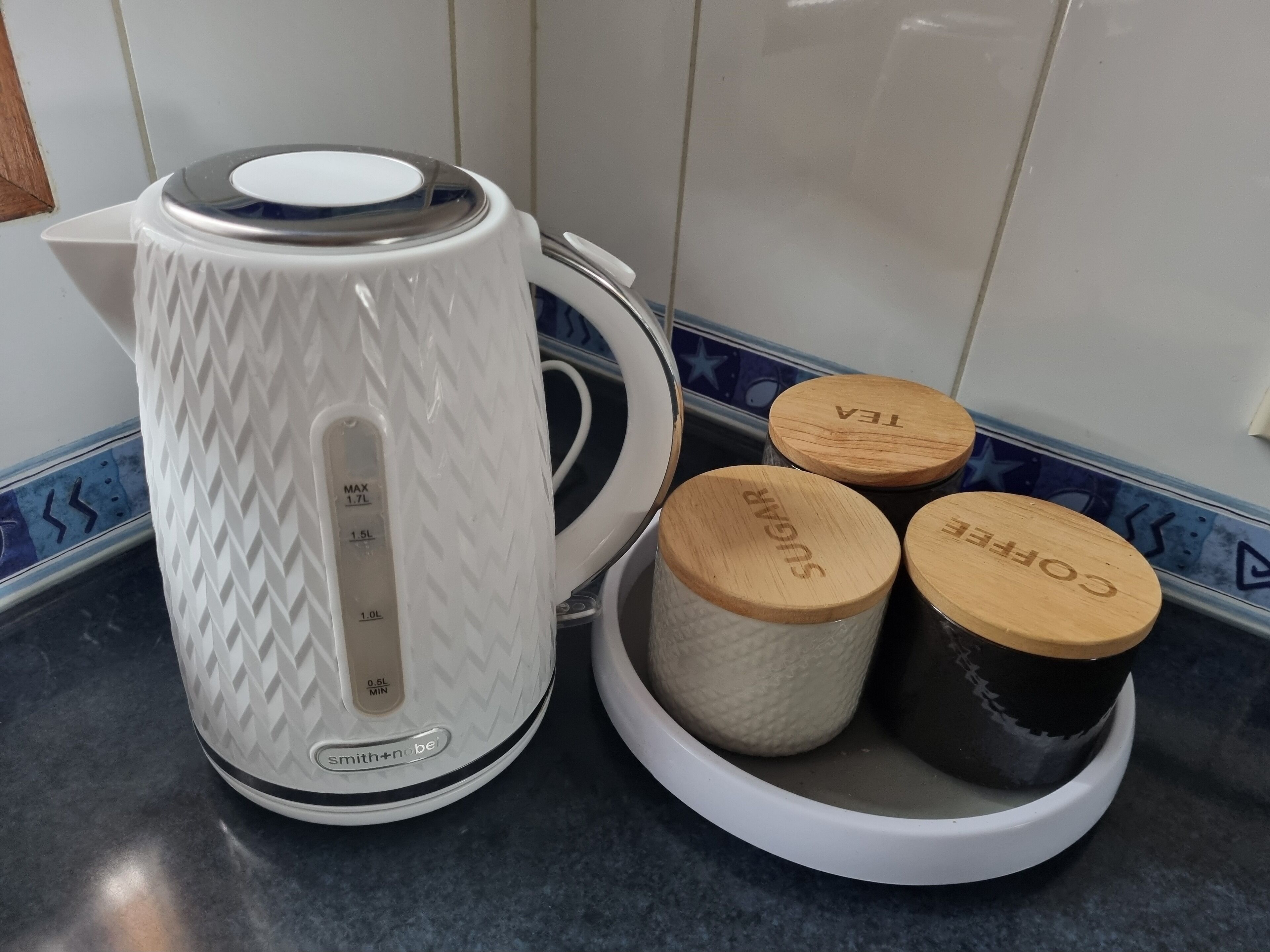 A white kettle with measurement markings and a container with three wooden lids labeled "SUGAR," "TEA," and "COFFEE."