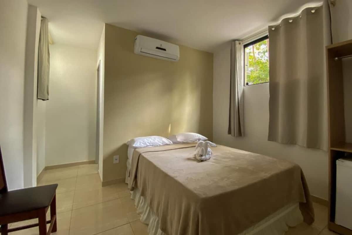 A bedroom with a bed, a chair, a window with curtains, an air conditioning unit, and a door.