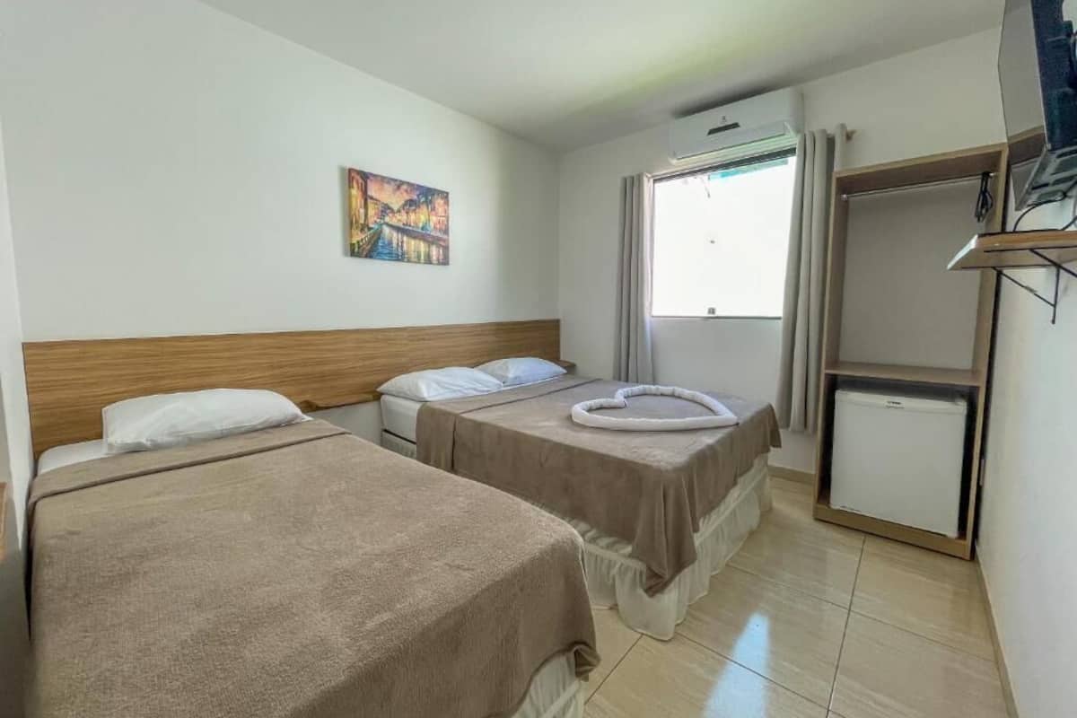 A hotel room with two beds, a small refrigerator, a painting on the wall, and a window with curtains.