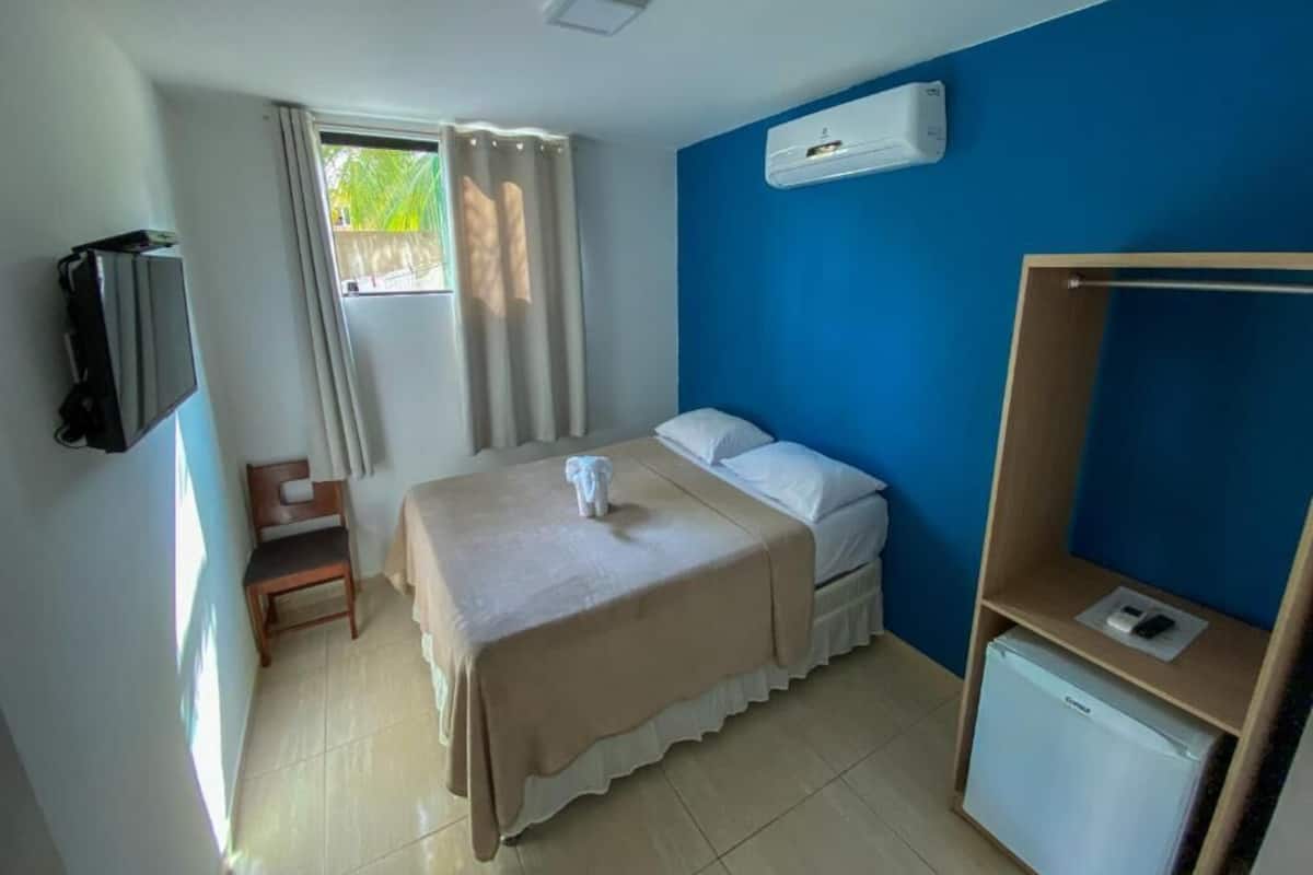 A hotel room with a bed, a desk, a chair, a TV, and an air conditioning unit.